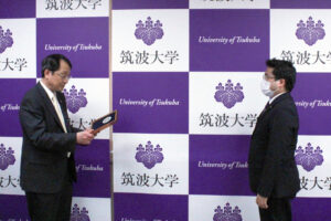 BEST FACULTY MEMBER表彰式の様子。写真左側に立った永田学長が表彰状を持ち、右側に立つ秋山先生に受け渡している